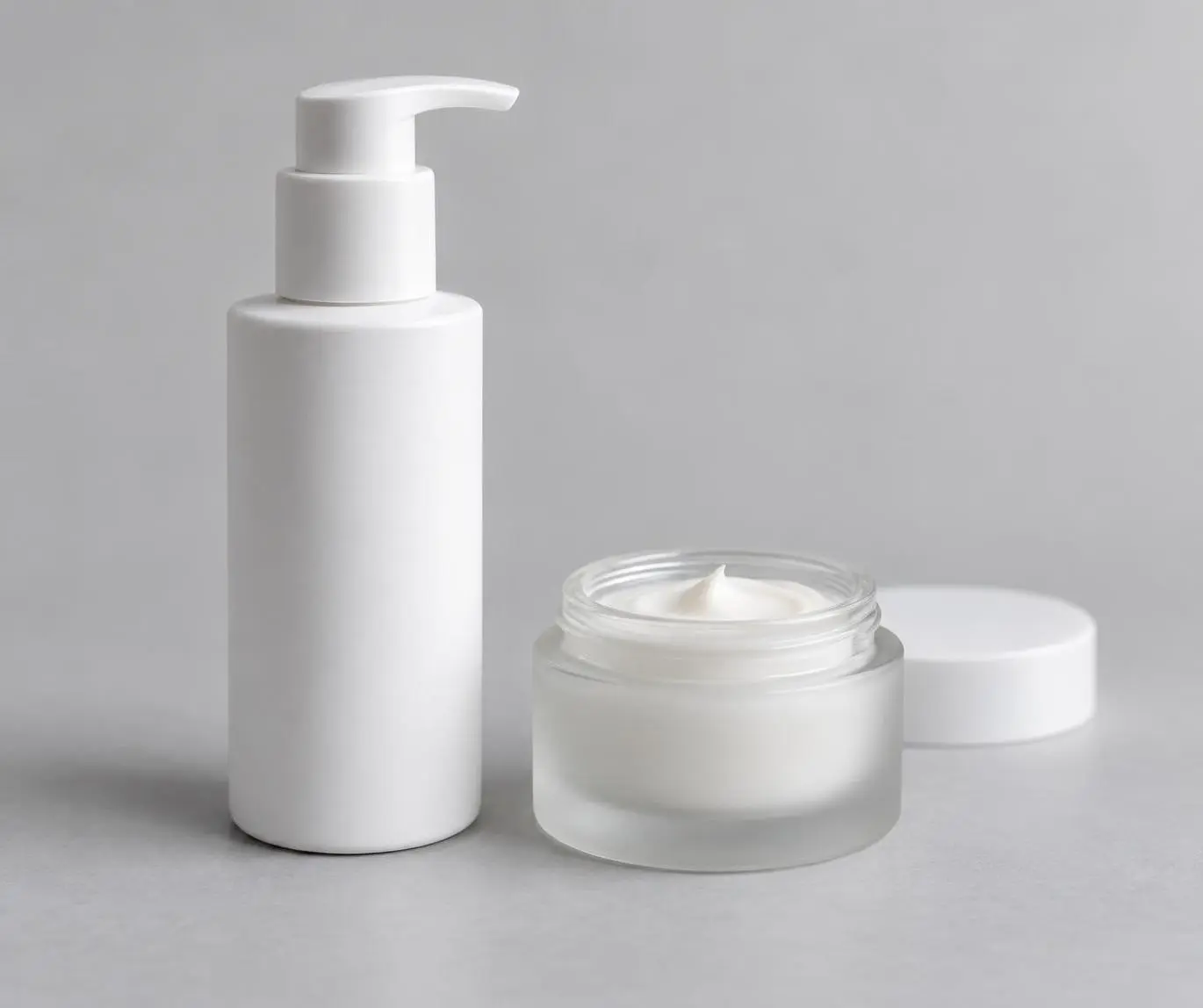 A plain pump bottle of face moisturizer beside a small jar of aftershave balm on a neutral grey background
