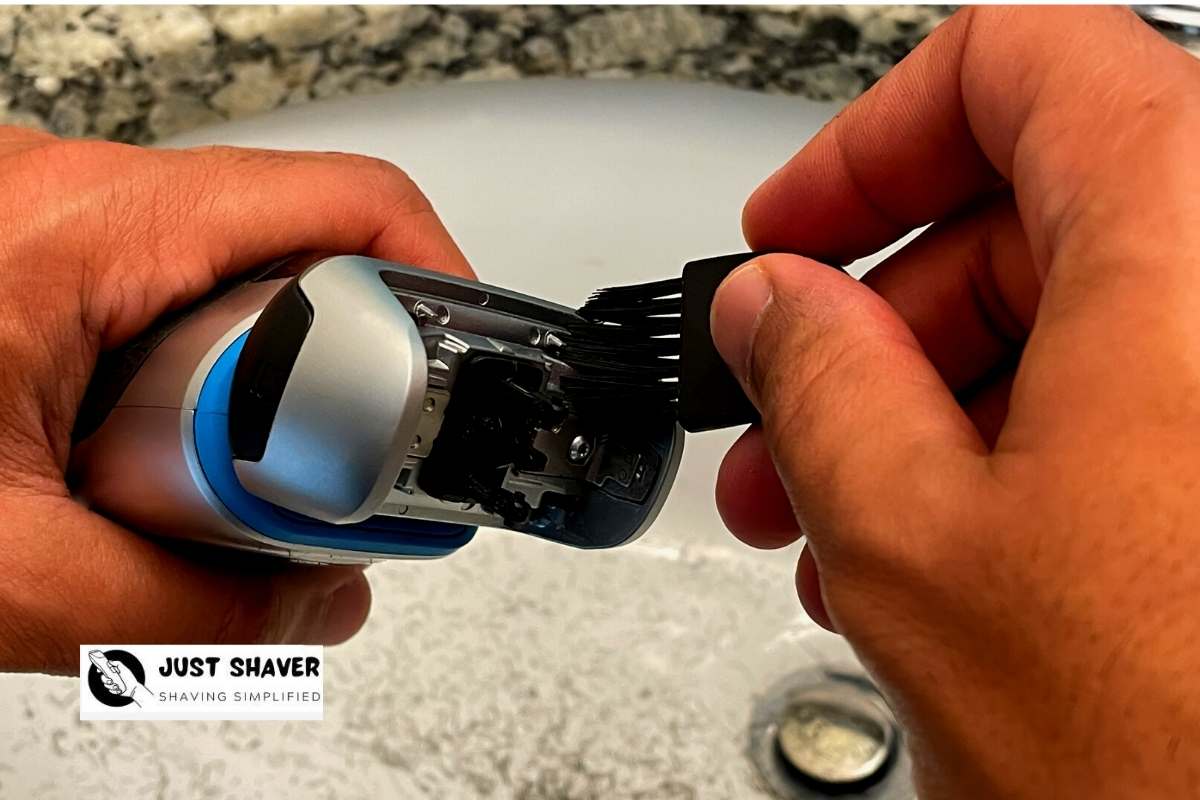 A hand brushing hair out of an open electric shaver head over a bathroom sink